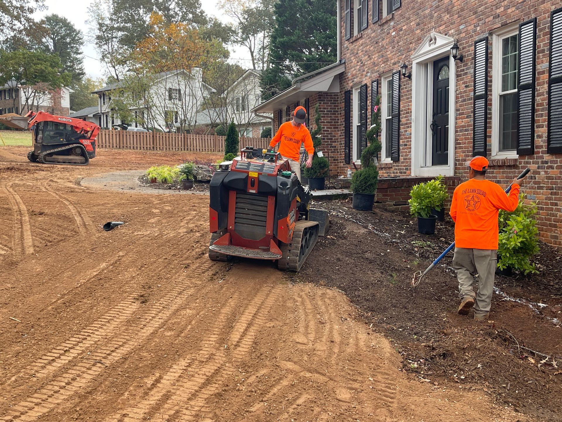 Two men are working on a dirt driveway in front of a brick house.