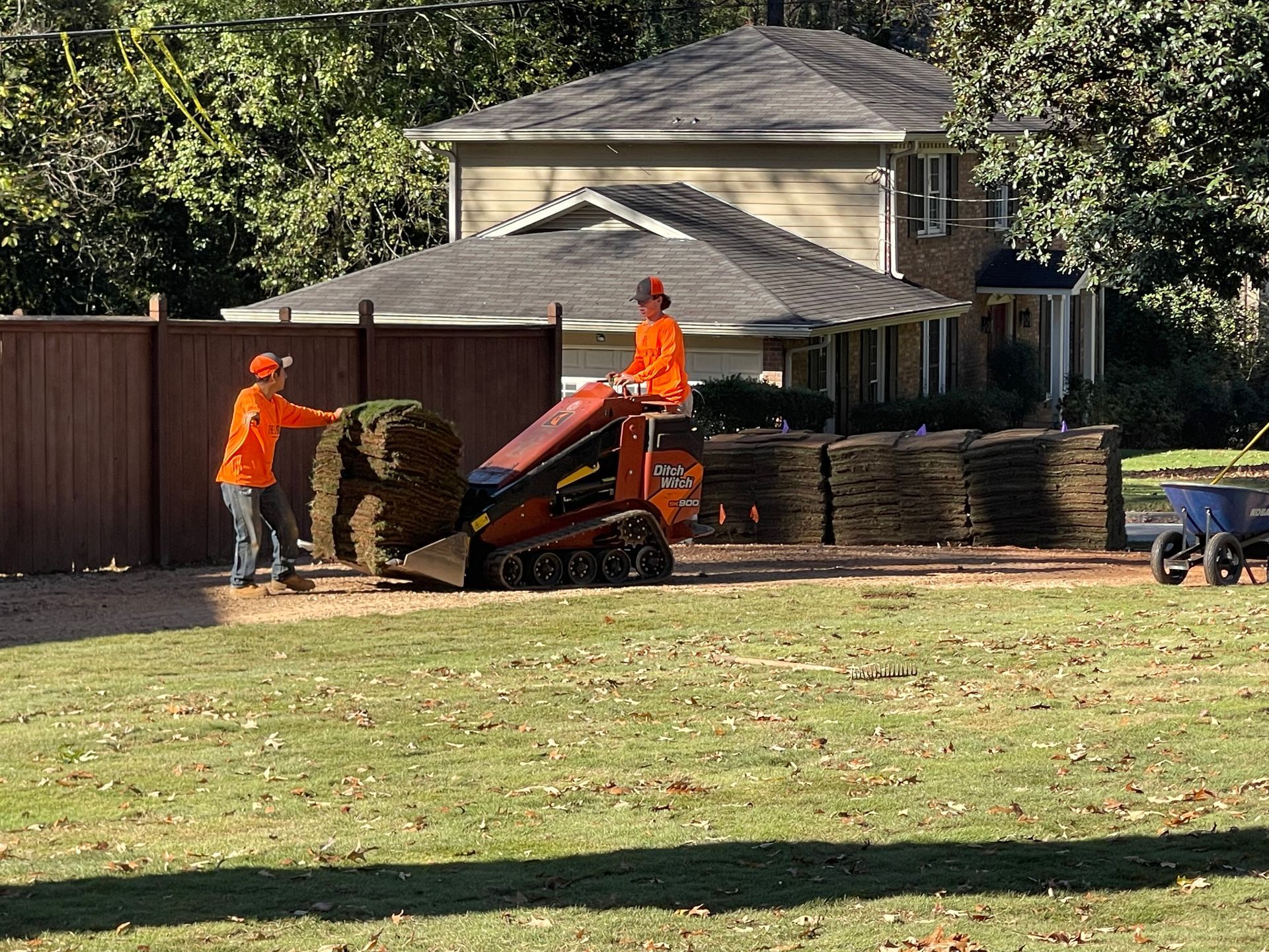 Two men are loading turf onto a tractor in a yard.