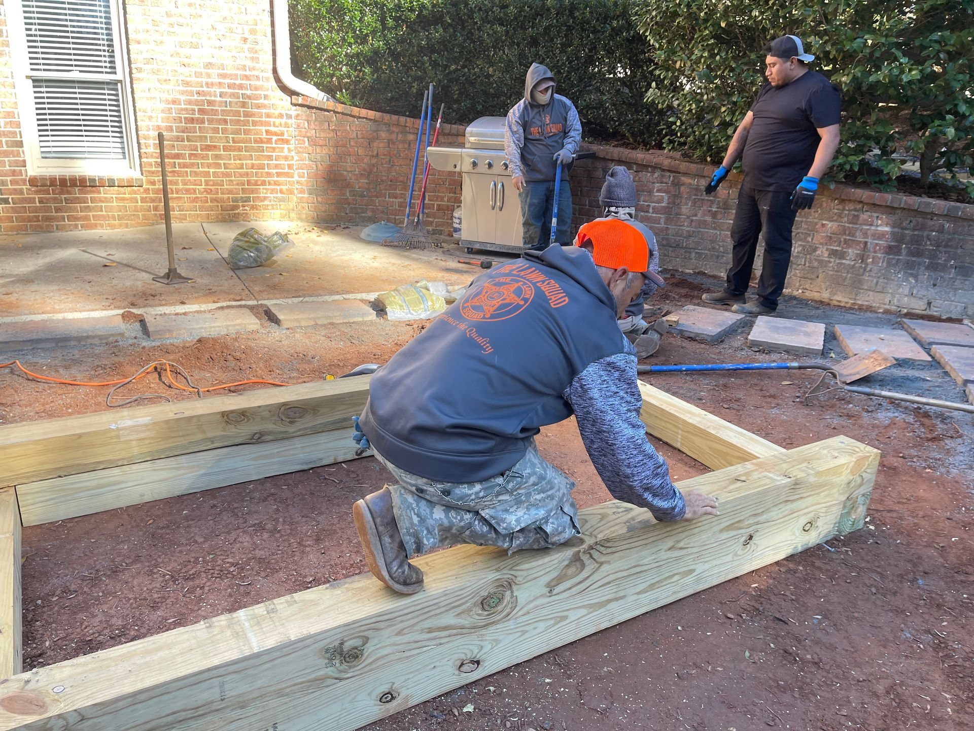 A man is kneeling on a wooden beam in a yard.