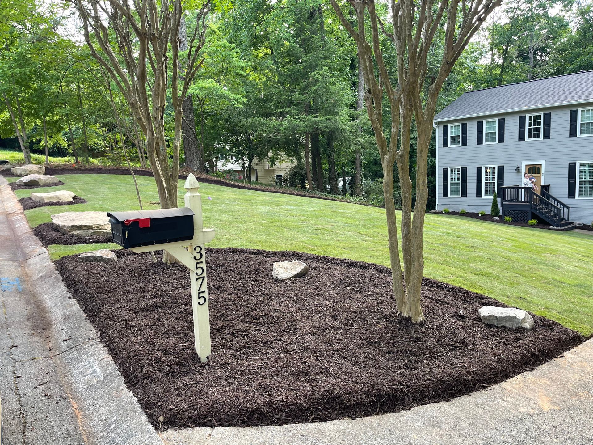 A mailbox is sitting in the middle of a lush green yard next to a house.