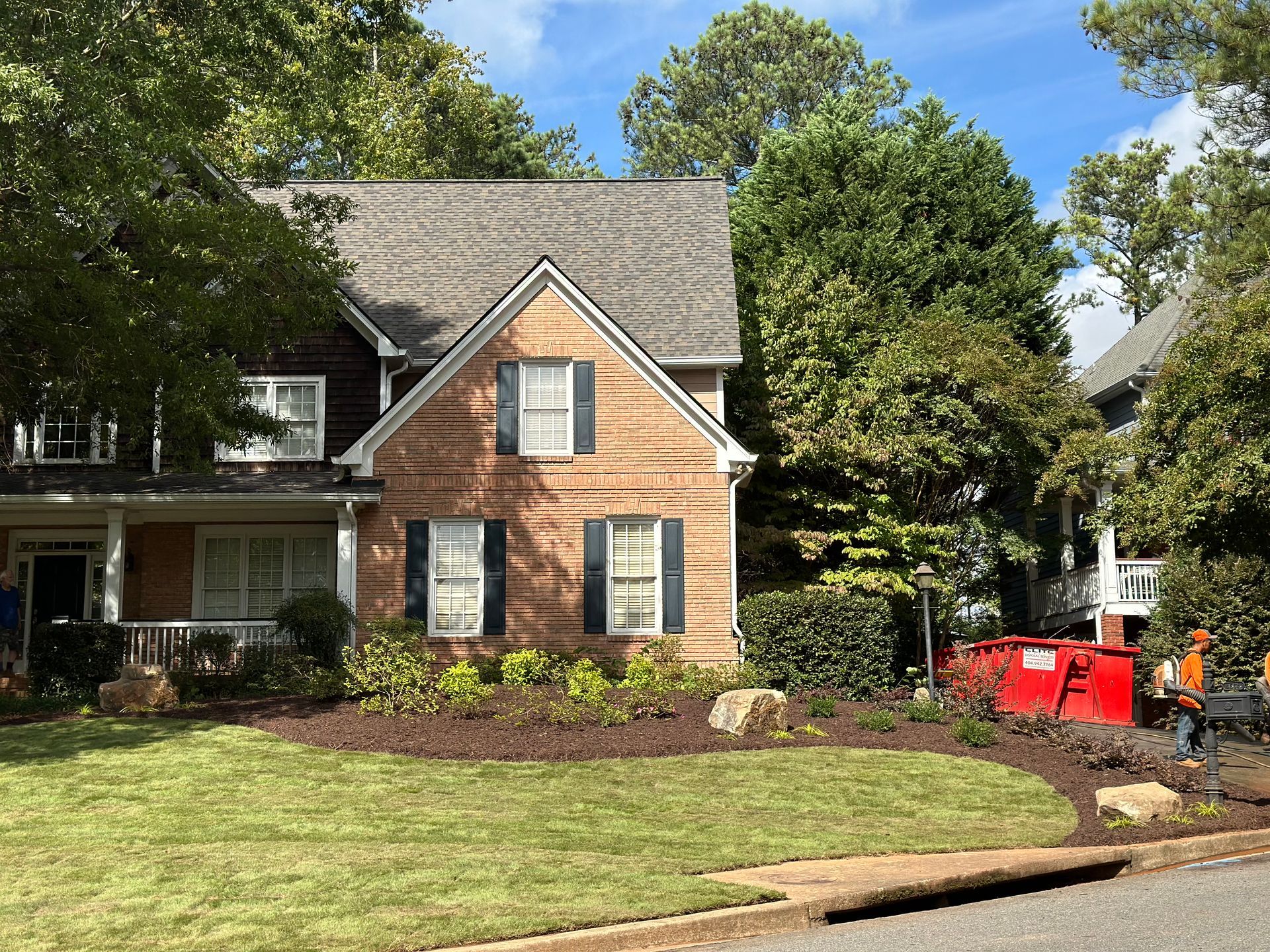 A large brick house with blue shutters and a red dumpster in front of it.
