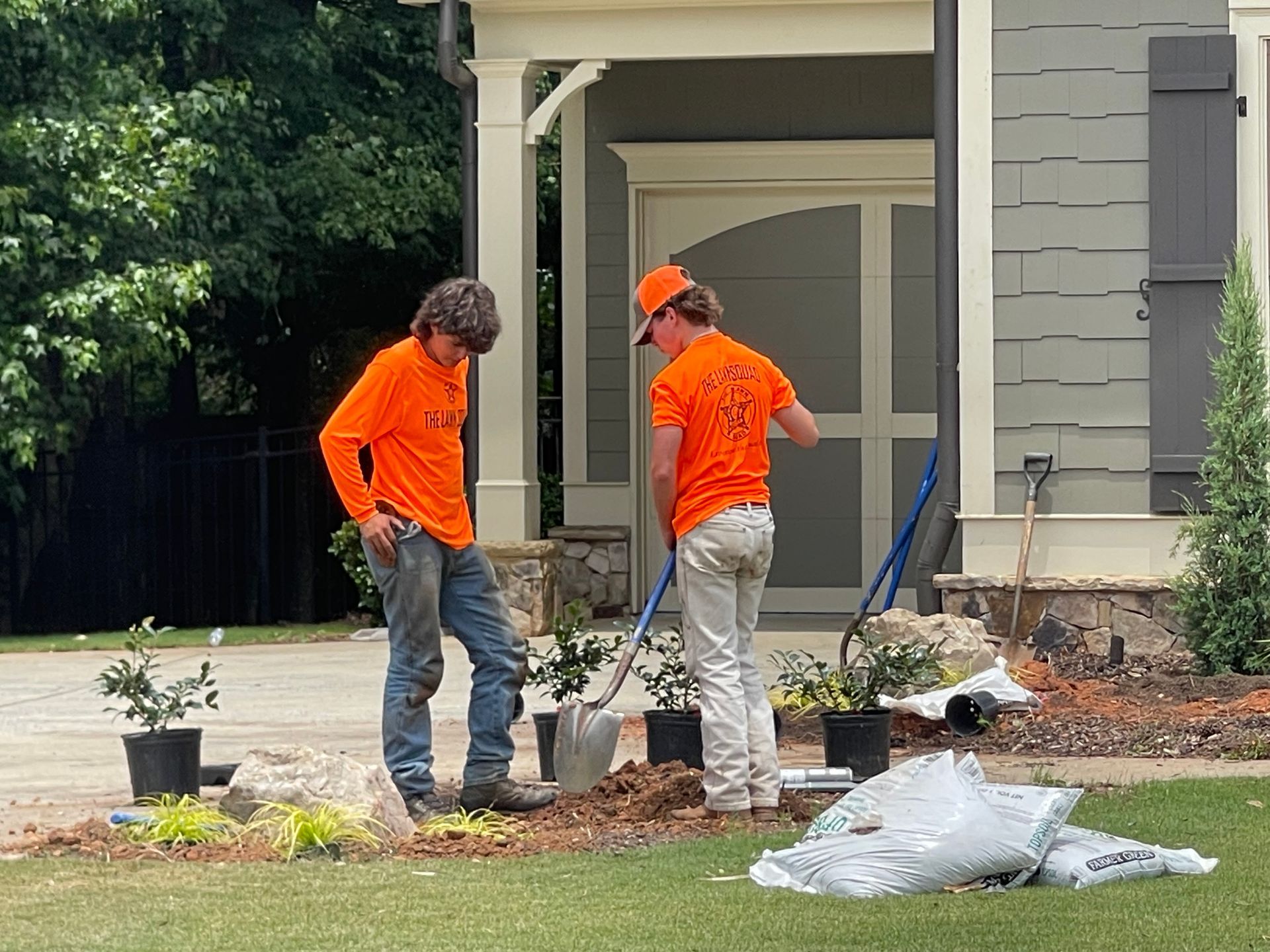 Two men in orange shirts are working on a lawn in front of a house.