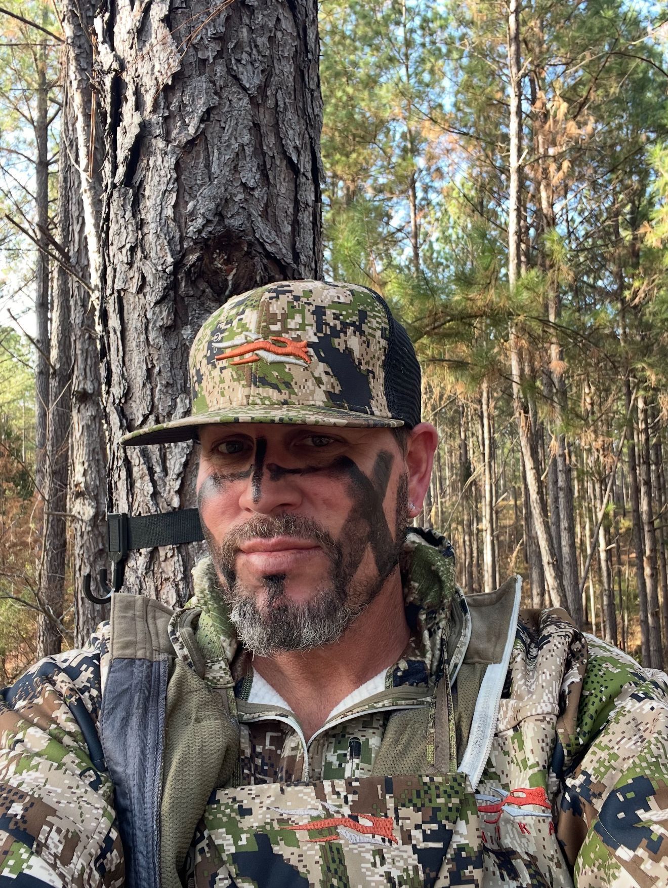 Man in camouflage, face paint, and hat; in a wooded area, likely hunting.