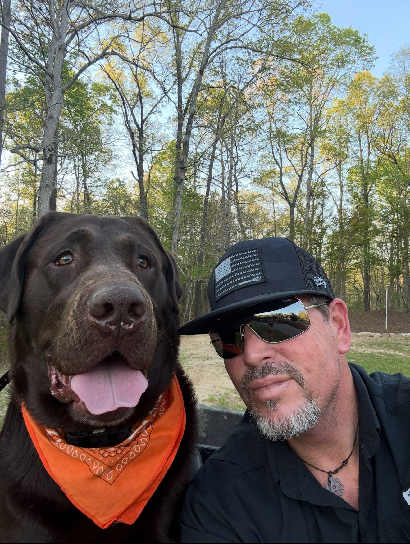 Man with sunglasses and dog wearing an orange bandana, smiling outdoors. Trees in background.