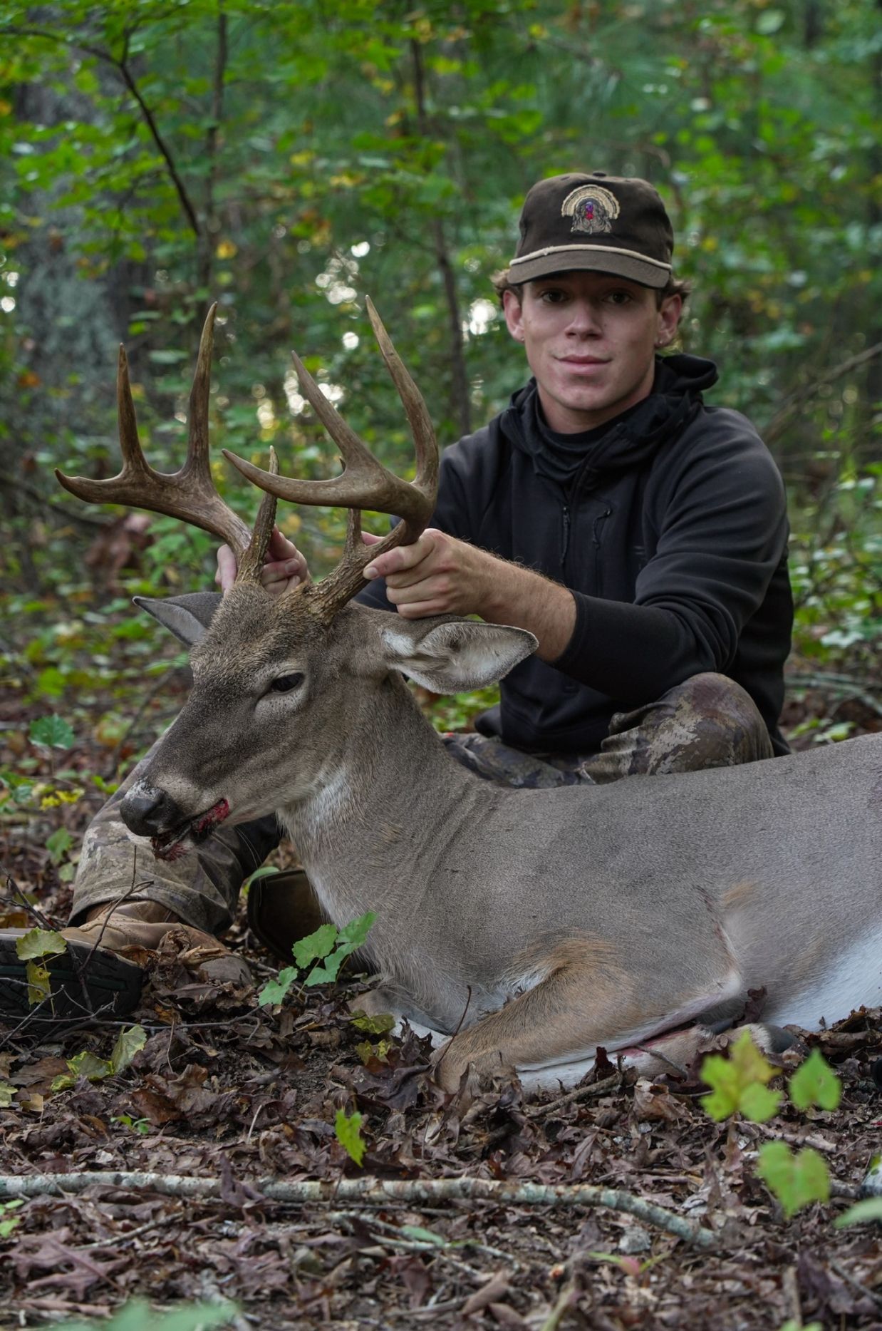 Man in a cap smiles next to a large deer in the woods. Deer is laying down.