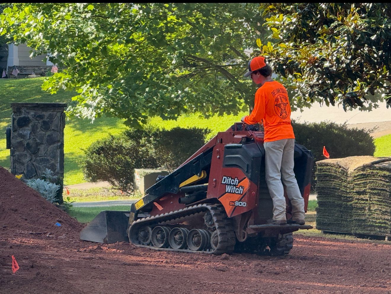 Person operating a small skid-steer loader on a dirt surface. Person is wearing orange. In front of a green backdrop.
