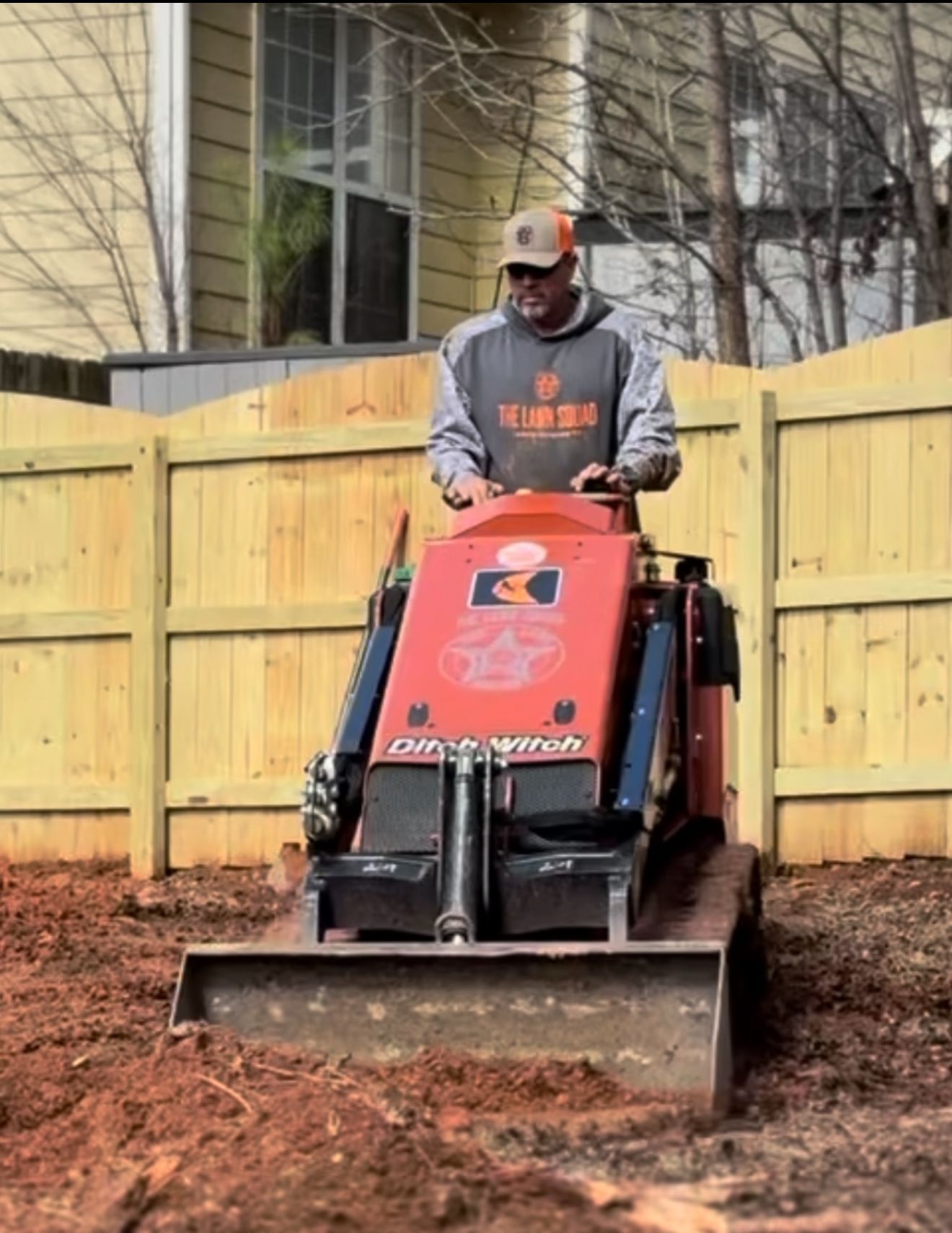 Man operating a small, orange ditch witch machine in a yard near a wooden fence.
