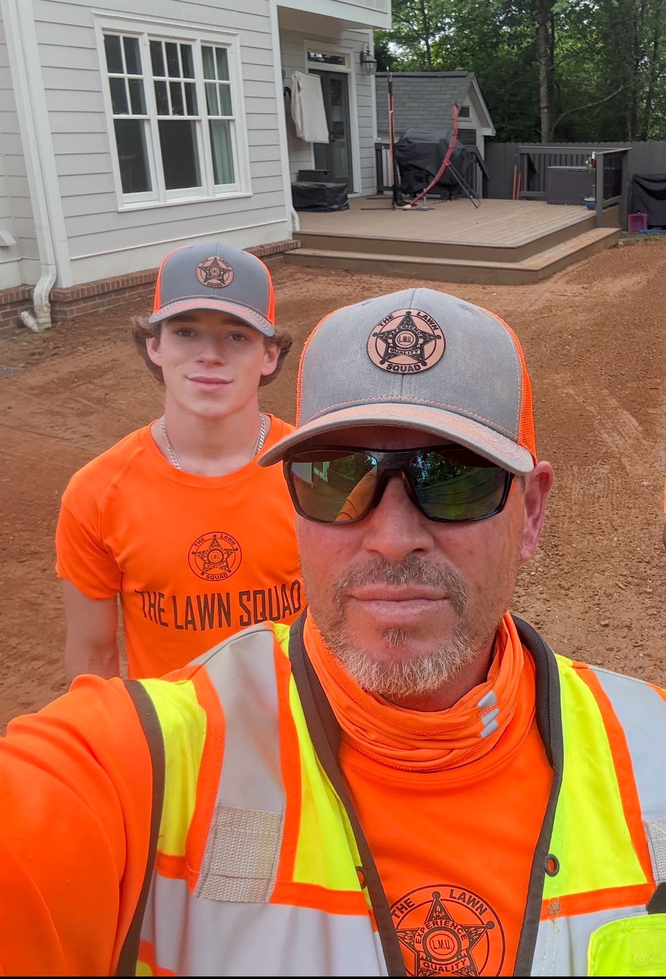 Two people wearing orange shirts and safety vests, standing outside a house.