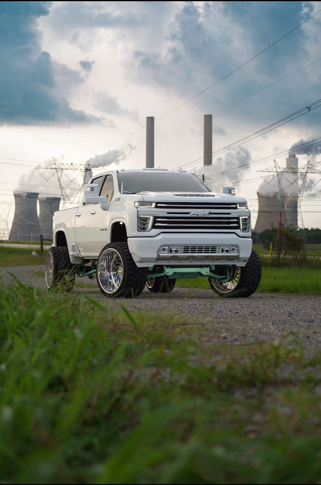 White lifted truck with chrome wheels, in front of a power plant, with green trim and exhaust stacks.