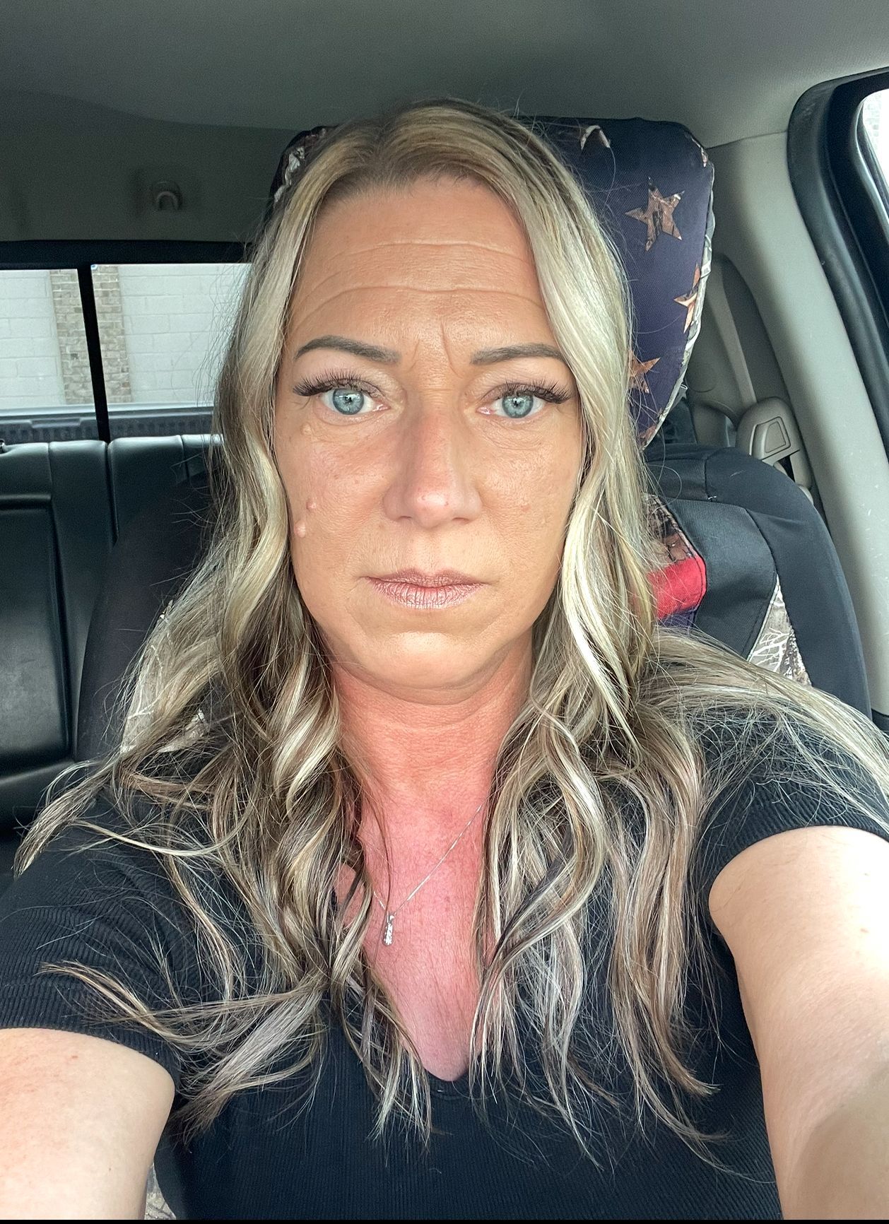 Woman with blonde and gray hair in a car. She has a neutral expression and wears a black shirt.