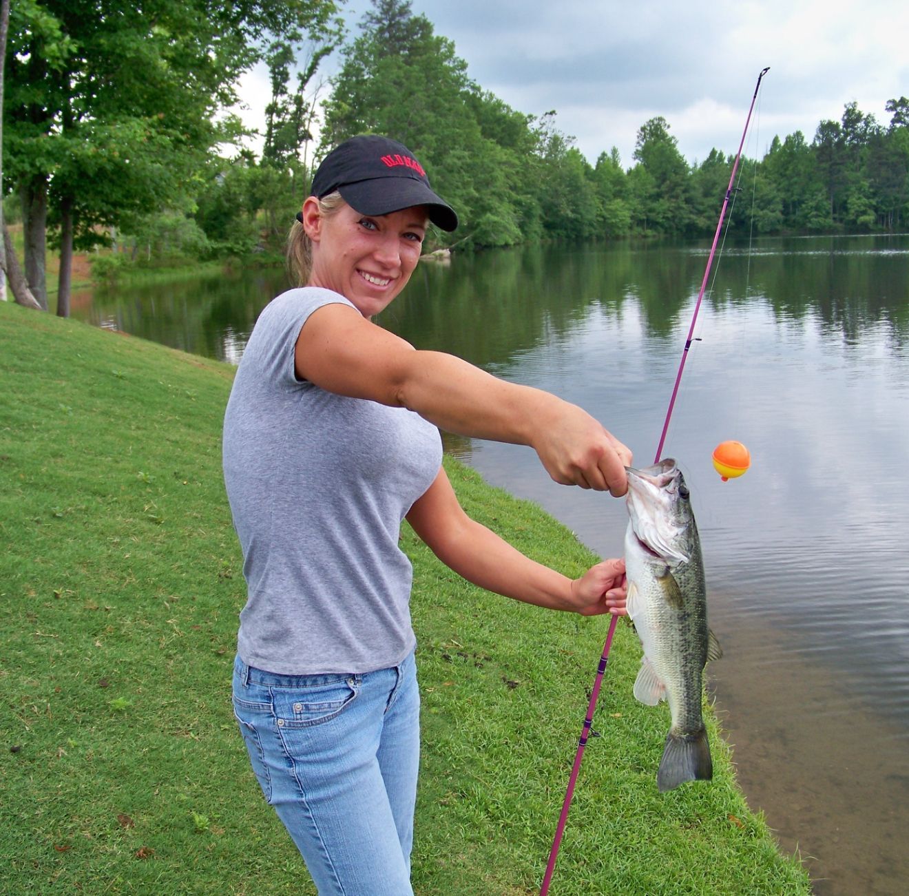 Woman holding a fish caught with a fishing rod by a lake, wearing a hat and smiling.