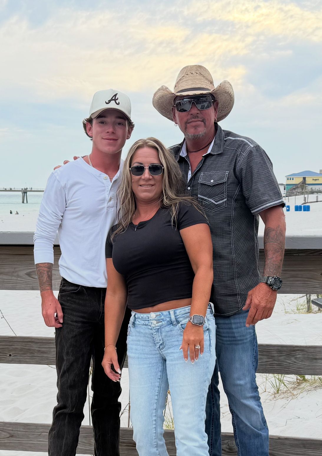 Family posing outdoors on wooden deck near beach; man in cowboy hat, woman in black top, younger man in baseball cap.