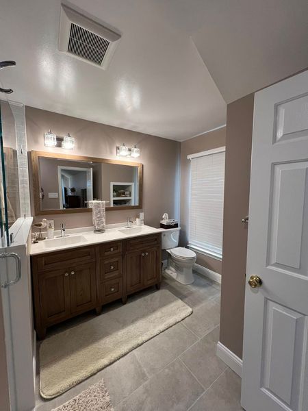 Bathroom with double vanity, toilet, and shower. Tan walls, gray tile, and wooden cabinets.