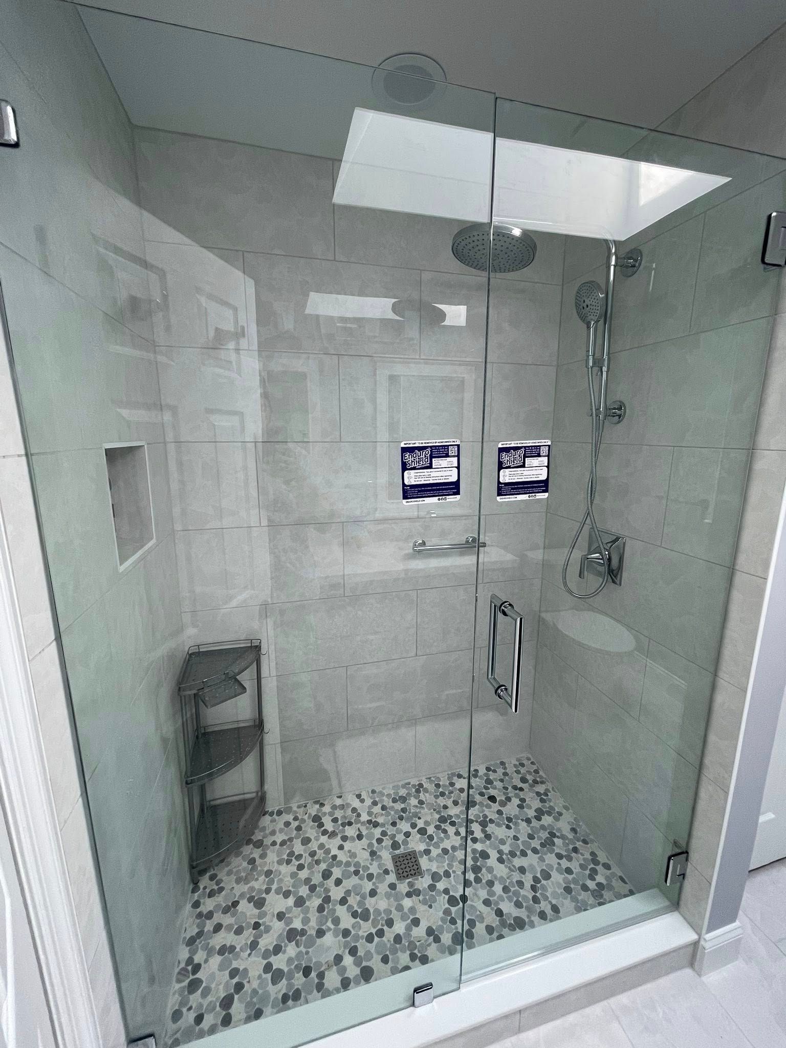 Shower with glass enclosure, pebble floor, gray tiled walls, rain showerhead, built-in shelving.