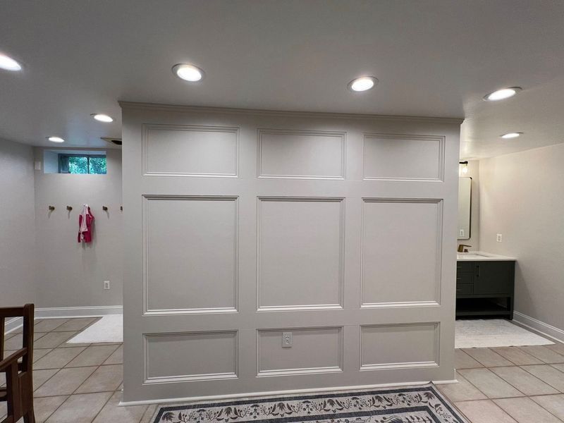 A white paneled wall divides a room with tile flooring and recessed lighting. A bathroom is visible on the right.