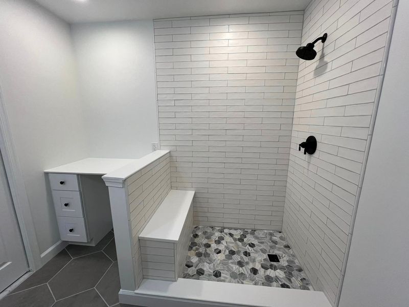 Bathroom with white brick tile shower, bench, and vanity with drawers. Dark fixtures and gray floor.