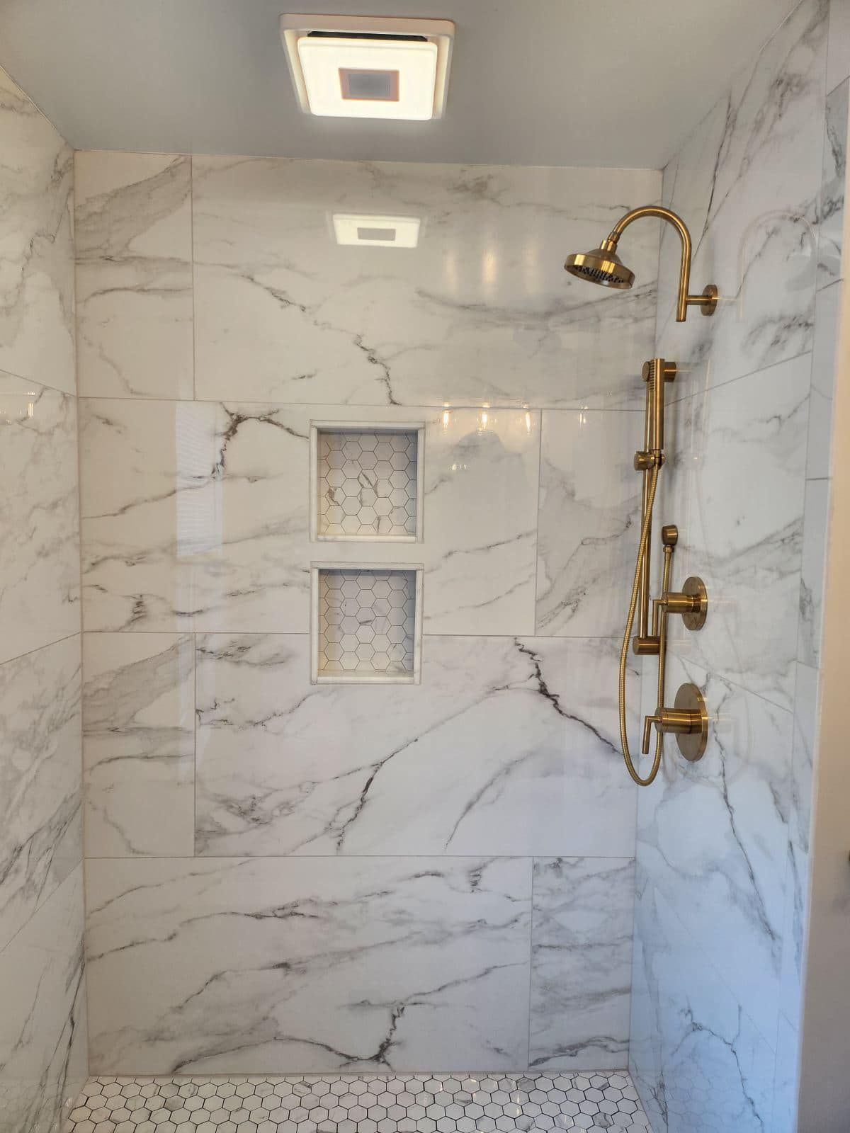 Marble-tiled shower with gold fixtures, two niches, and a pebble-stone floor. Overhead lighting in a modern bathroom.