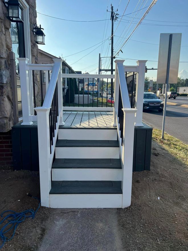 White and green deck with stairs, black railing, leading to a business. Road and cars in background.