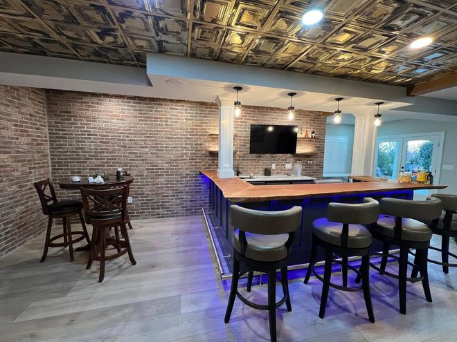 Basement bar with brick accent wall, ornate ceiling, and wooden bar with stools.
