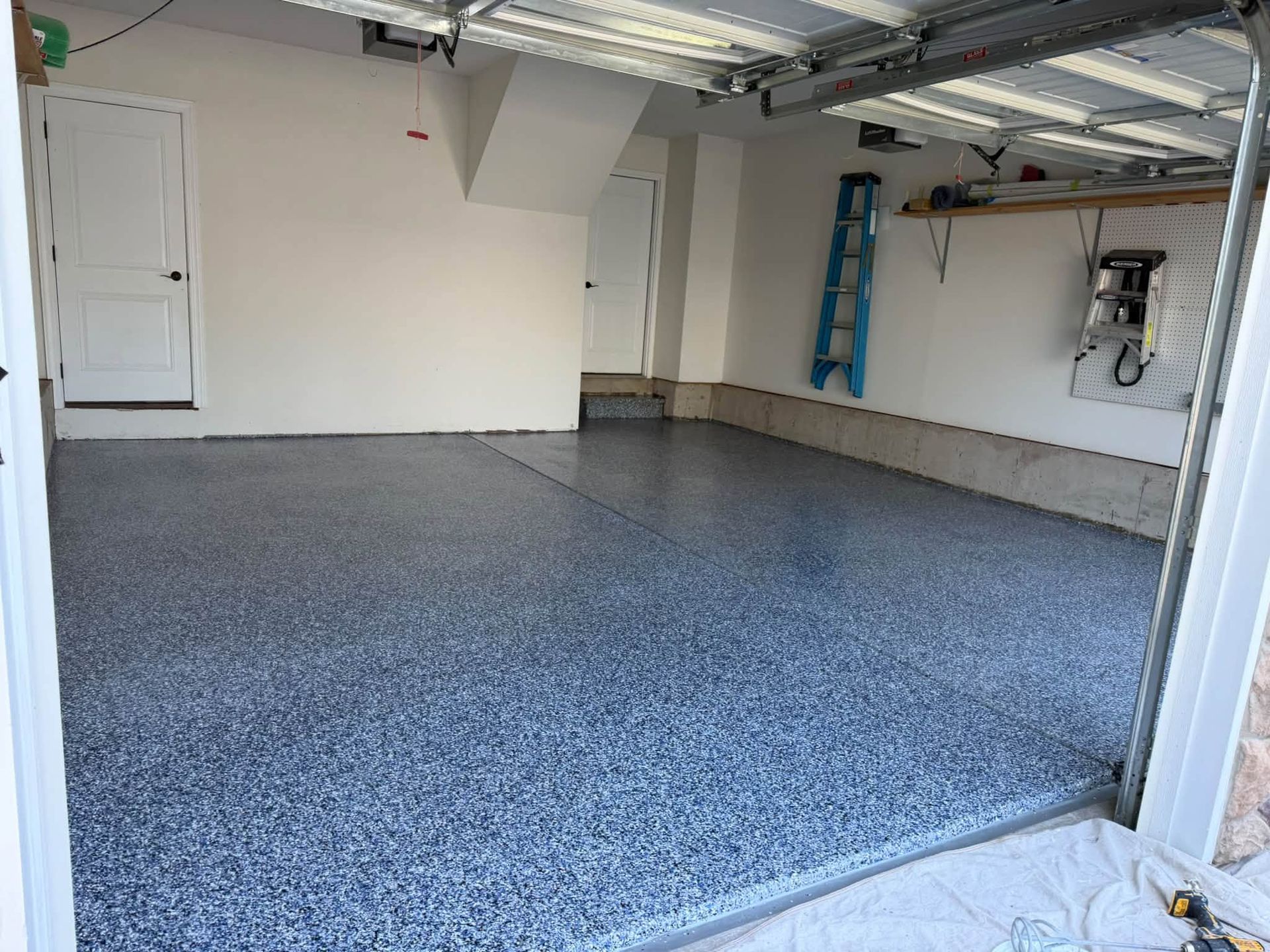 Empty garage interior with speckled gray epoxy floor, white walls, and a blue ladder near the back wall