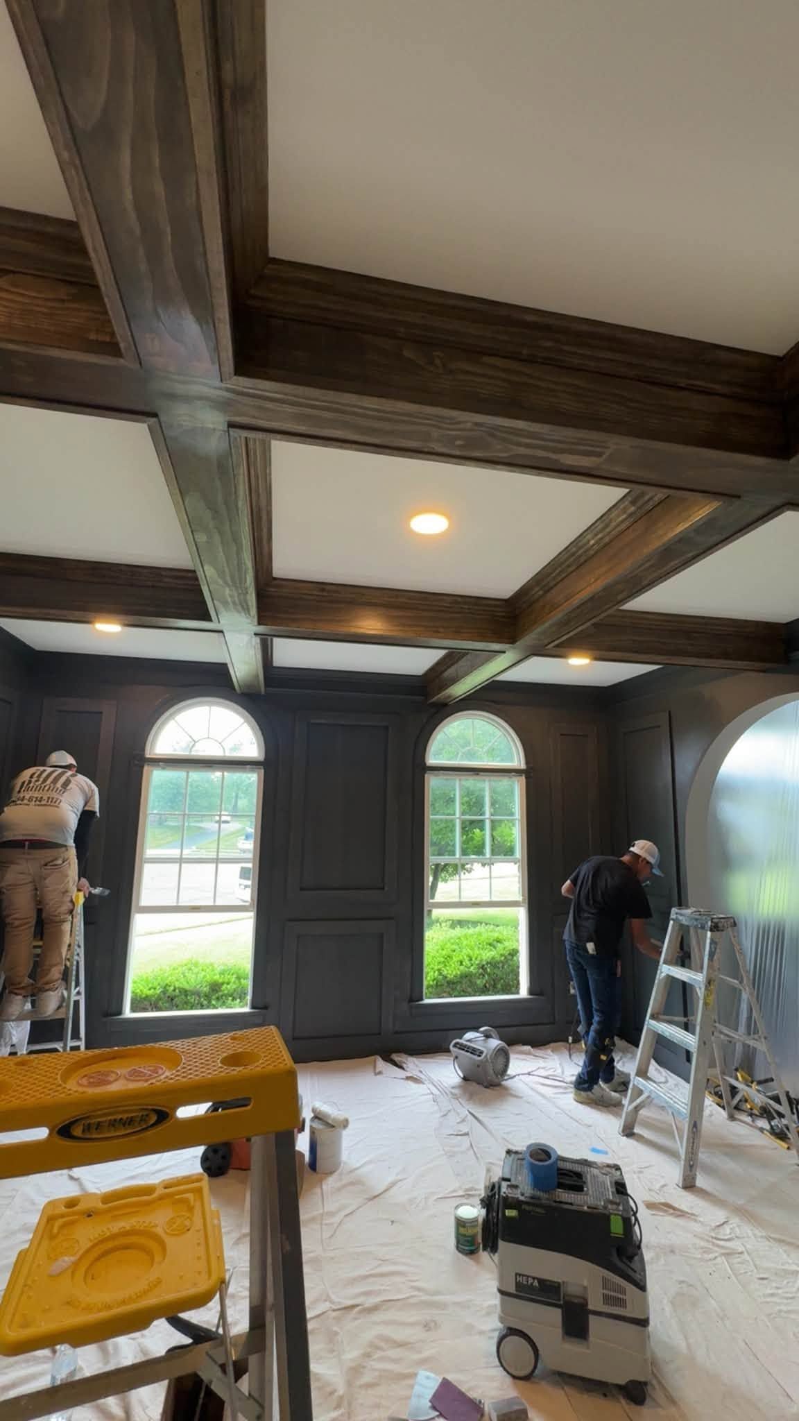 Workers renovating a room with dark wood beams, arched windows, ladders, and tools on the floor.