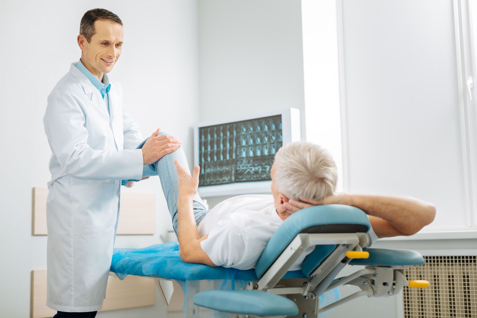 Doctor examining a patient’s leg in a bright clinic room with an X-ray display