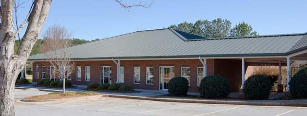 Single-story brick building with a green metal roof and covered walkway, framed by trees under a blue sky.