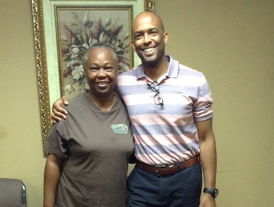 Two people smiling and standing together in an office, with framed wall art behind them.