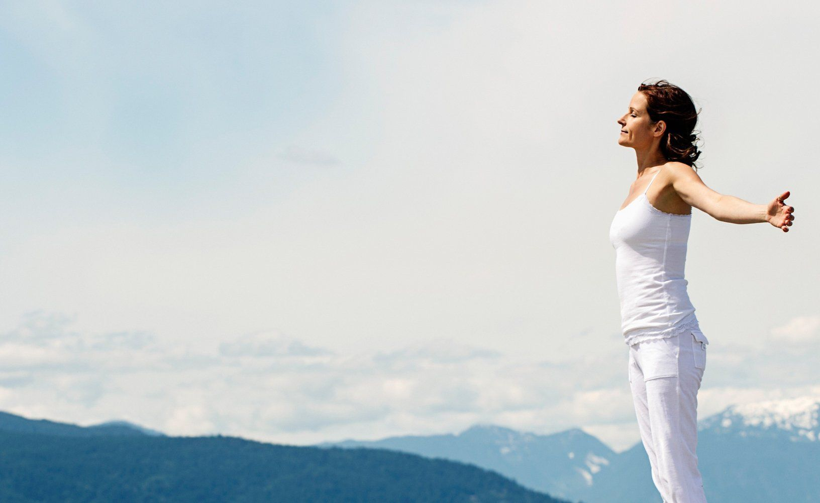 Person in white stands with arms outstretched on a mountain overlooking a cloudy sky.