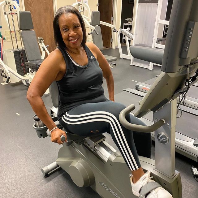 Woman smiling on an exercise machine in a gym, wearing a black tank top and striped leggings.