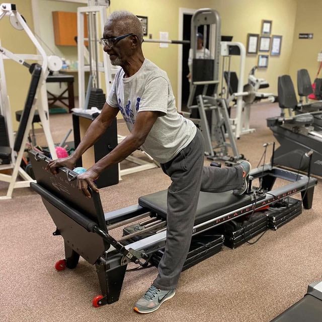 Man exercising on a Pilates reformer in a gym, stretching one leg back while holding the frame.