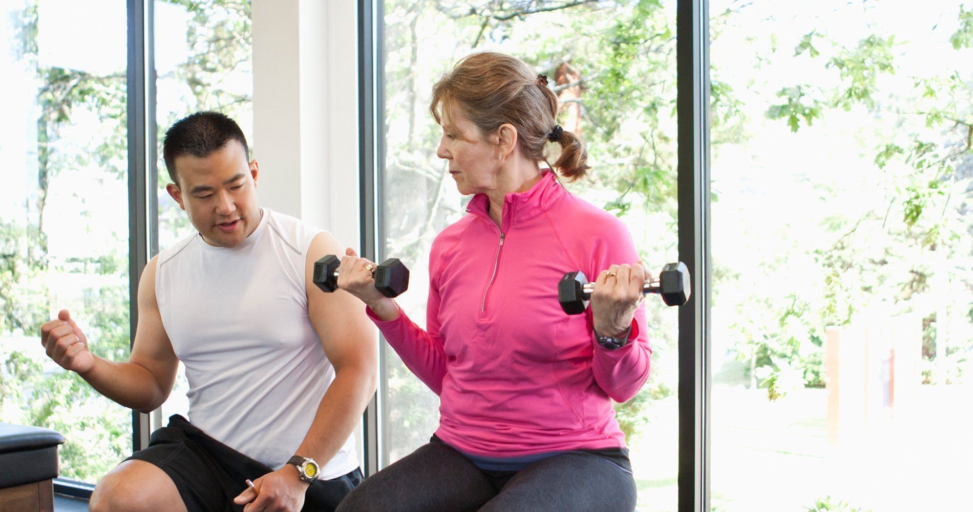 Two people lifting dumbbells in a bright gym, seated by a large window.
