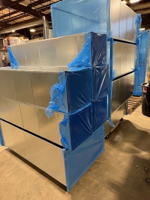 A stack of rectangular metal HVAC duct sections wrapped in protective blue plastic film, sitting on a warehouse floor.