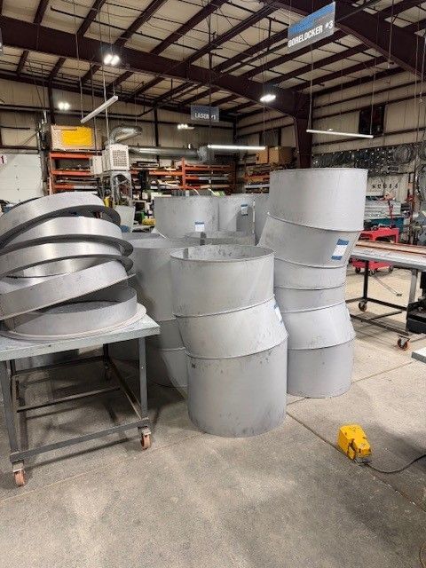 Stacked metal cylindrical duct segments in a workshop setting, with some pieces on a metal rolling cart.