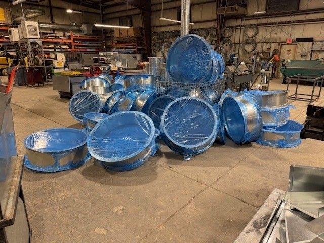 A large pile of circular metal duct connectors wrapped in blue protective plastic on a workshop floor.