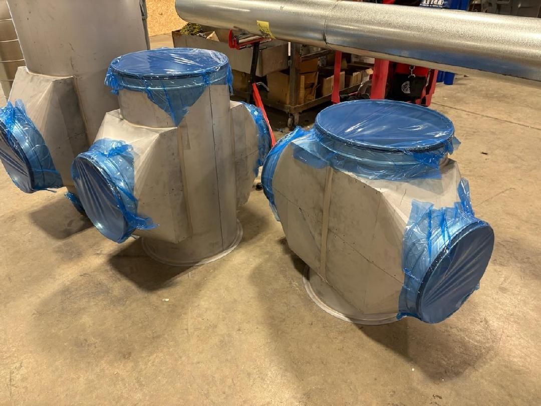 Industrial metal pipe components with blue protective covers on the circular openings, positioned on a workshop floor.