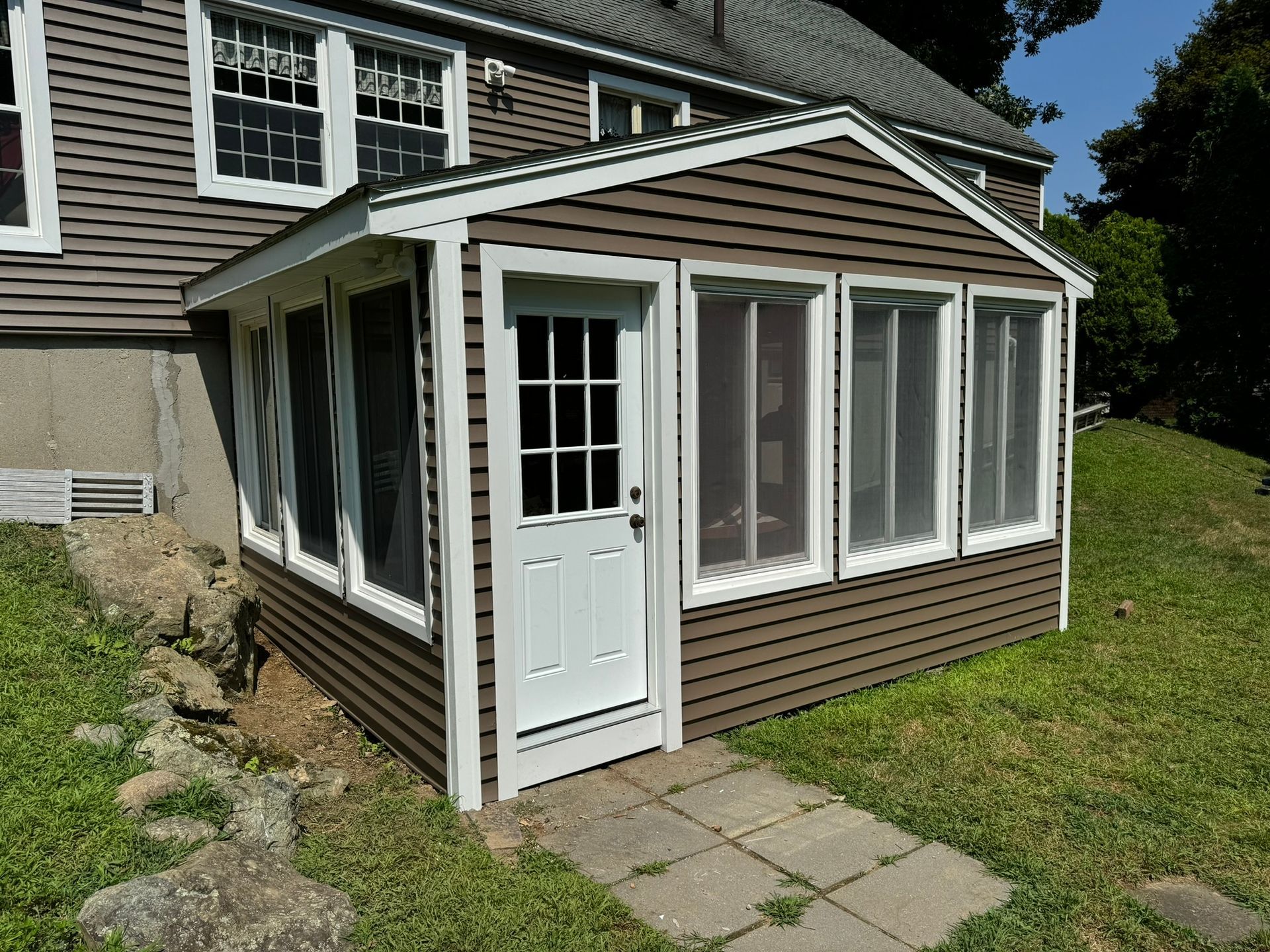 Small brown sunroom addition with white trim and door beside a house, with patio stones and grass.