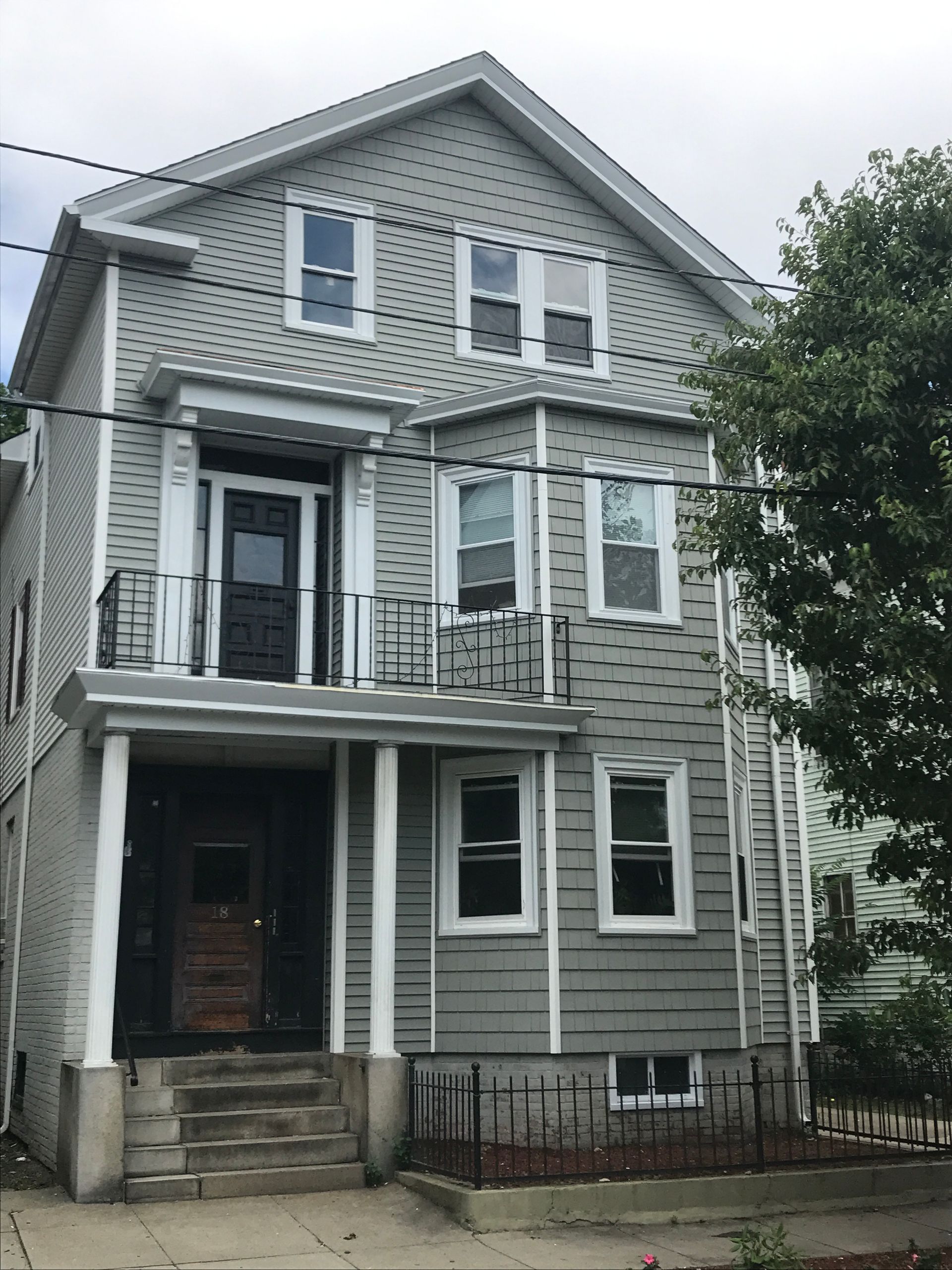 Three-story gray house with white trim, front porch, and small iron fence