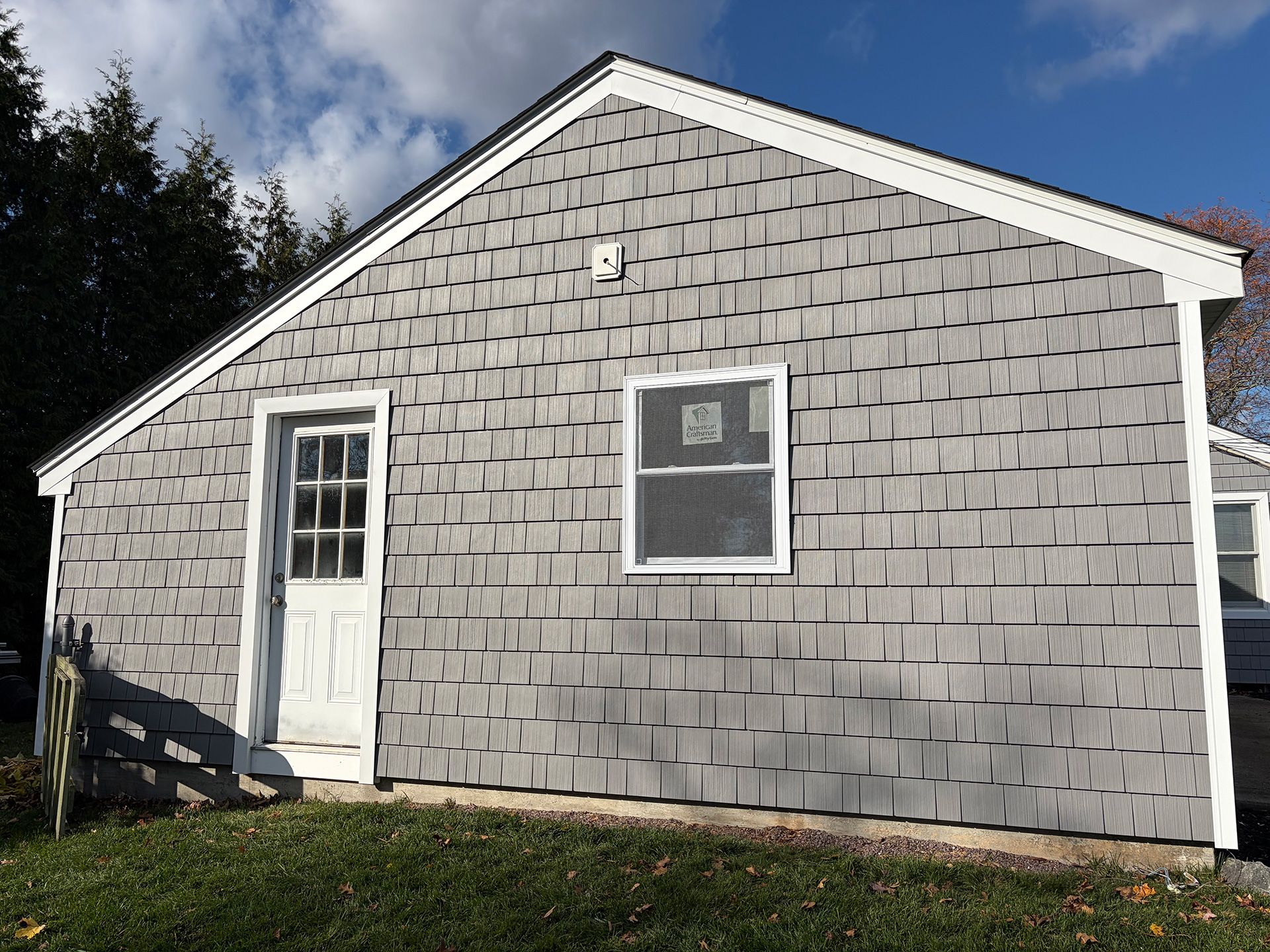 Small gray shingled garage or shed with a white door and window, viewed from outside on a sunny day