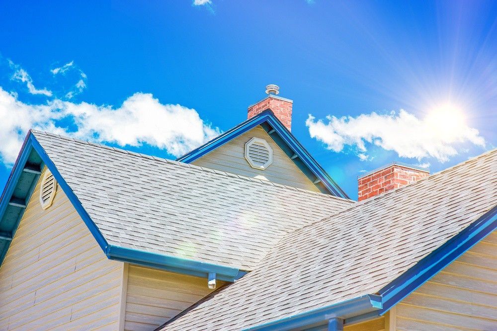 Bright blue sky above a house roof with white shingles and blue trim in sunlight