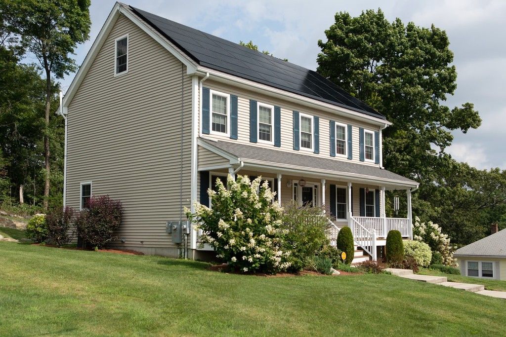 Suburban two-story beige house with white trim, front porch, and green lawn under a cloudy sky