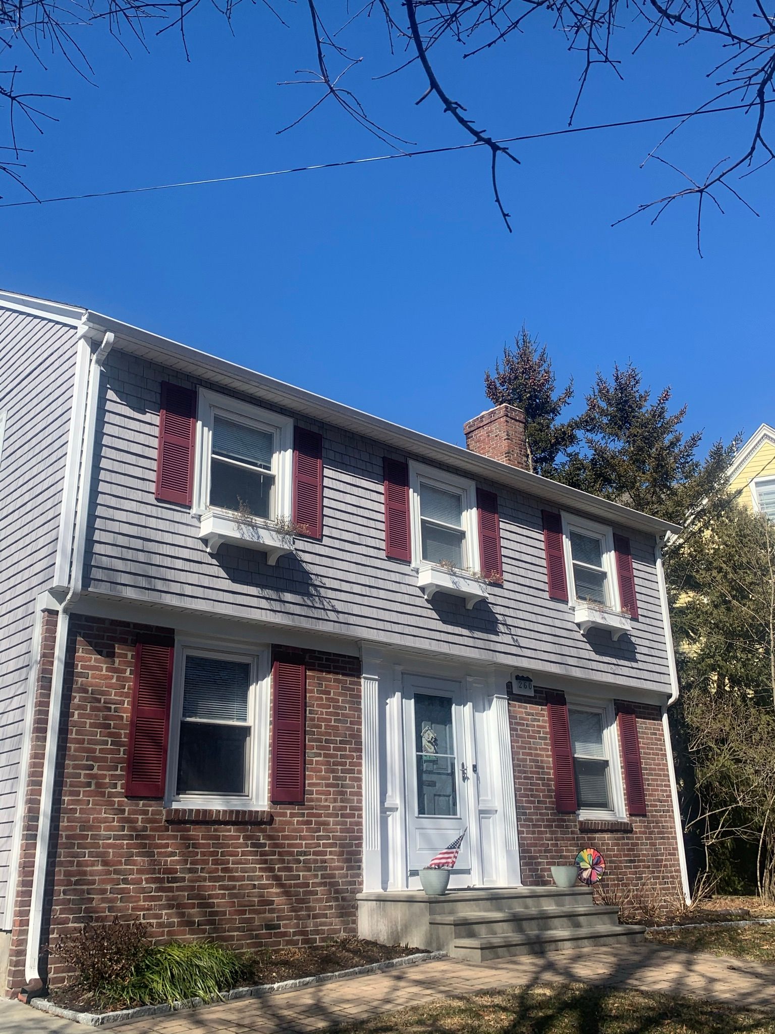 Two-story brick house with gray siding, red shutters, and a white front door under a clear blue sky.