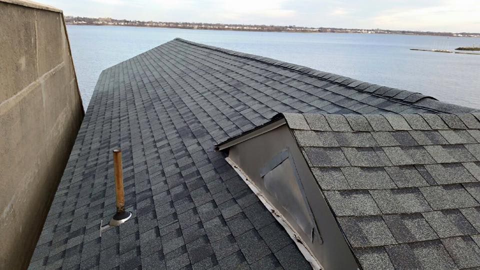 Shingled rooftop beside water, with a small skylight and vent pipe under a cloudy sky