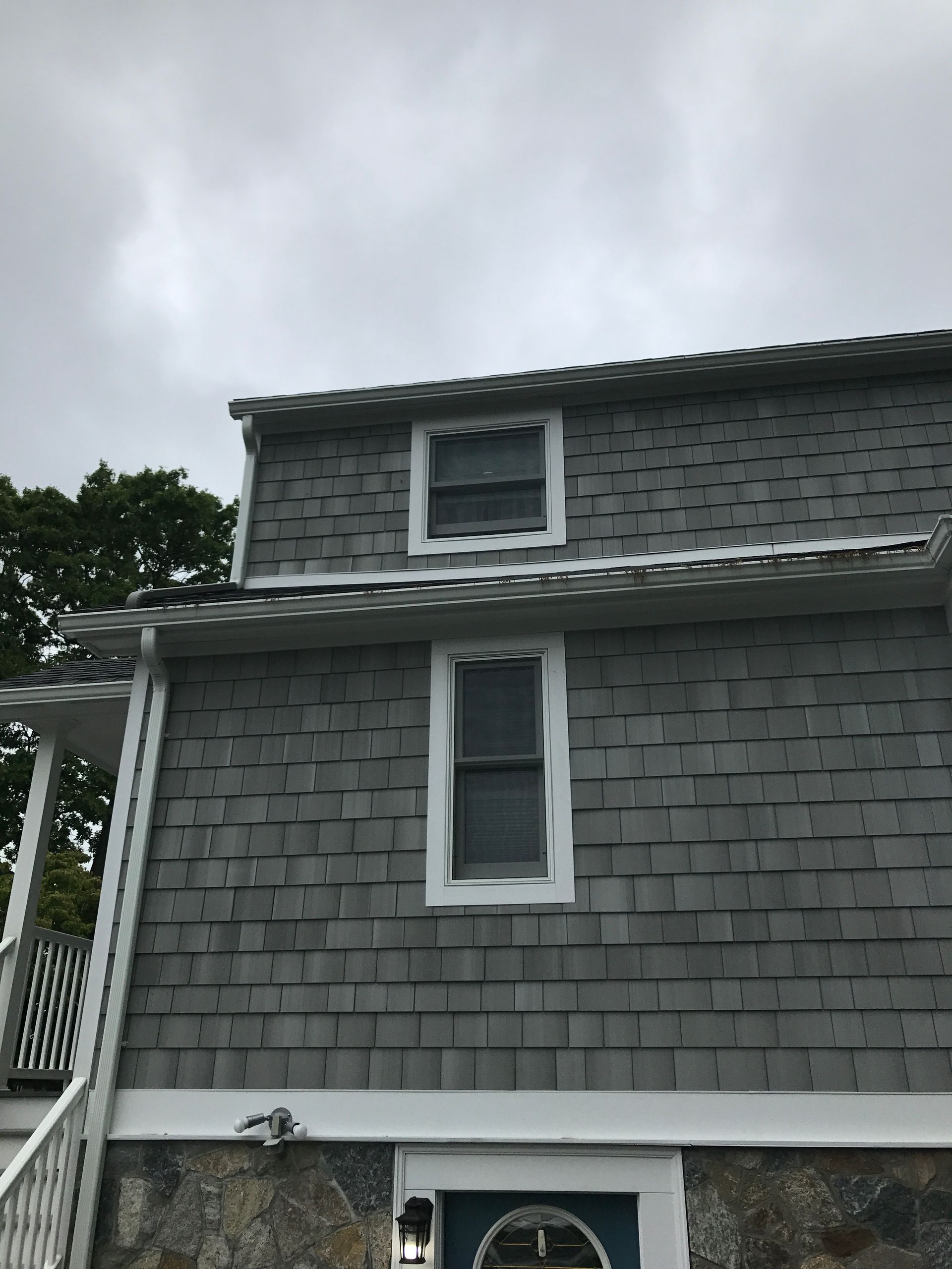 Gray shingled house exterior with white-trimmed windows and a porch roof under an overcast sky