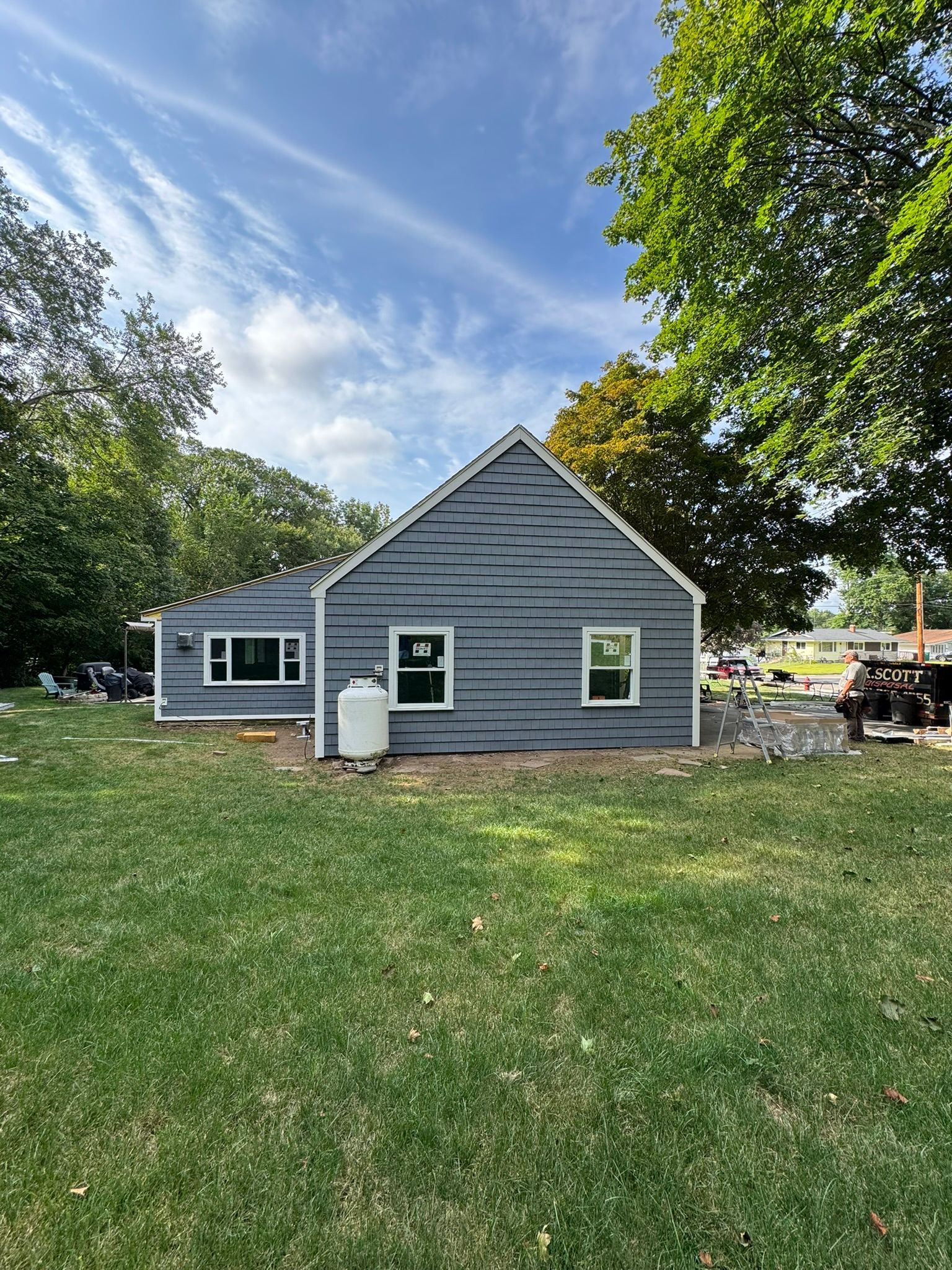 Gray house with white trim in a grassy yard, surrounded by trees under a blue sky