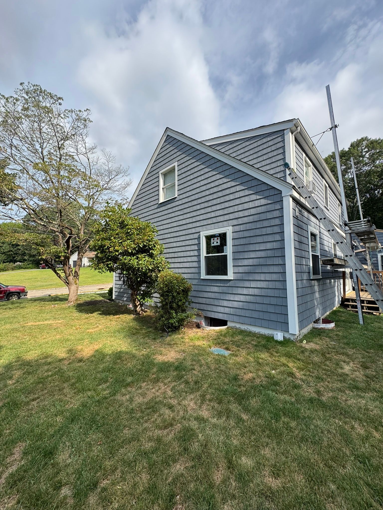 Gray house with white trim and outdoor stairs beside a grassy yard under a cloudy sky