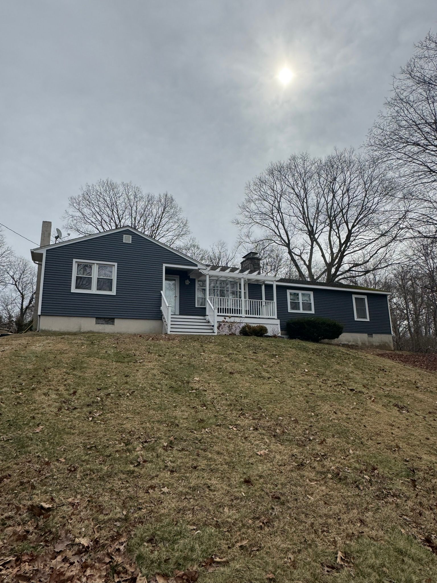 Blue house on a grassy hill under a cloudy sky, with a white front porch and leafless trees.