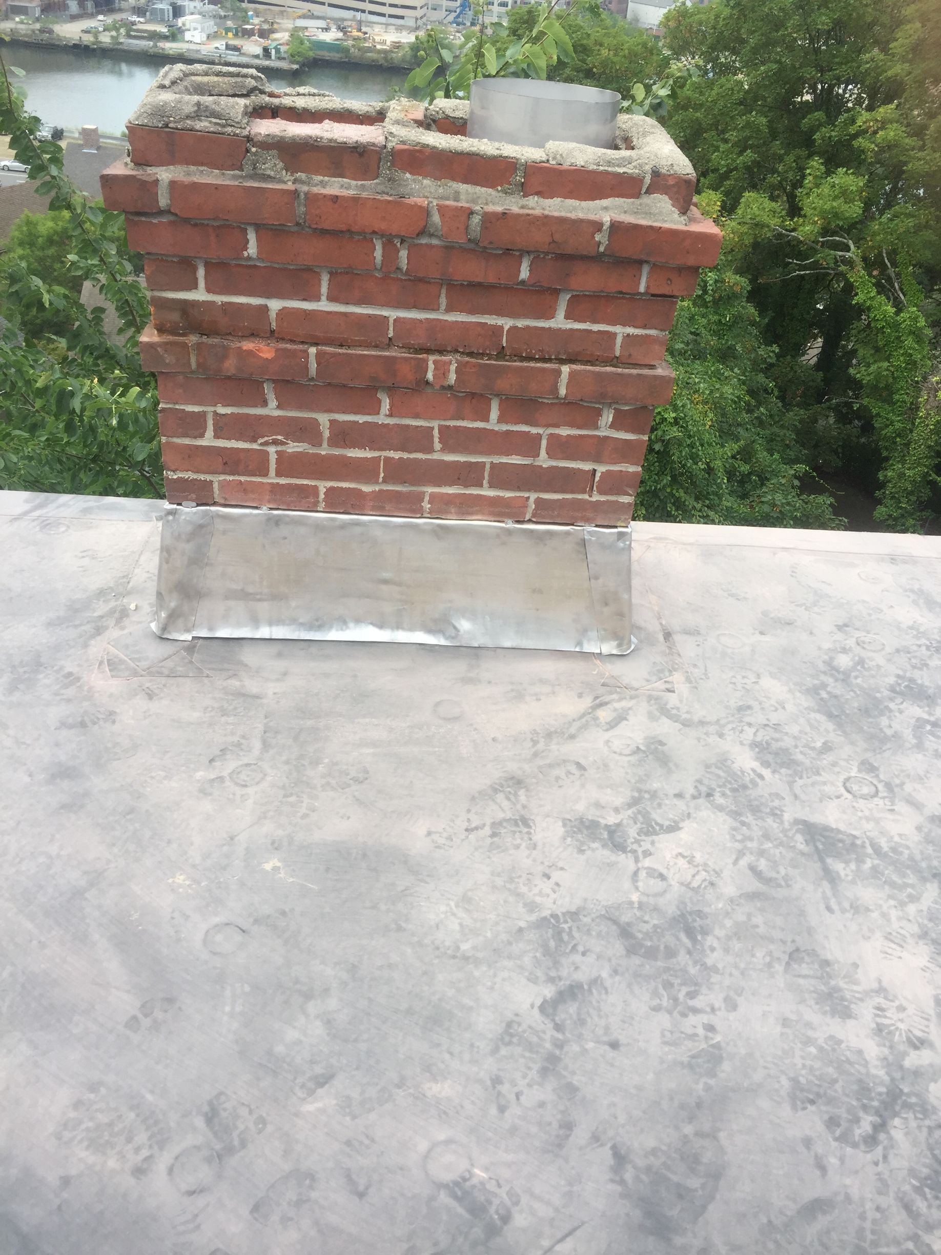 Red brick rooftop chimney with a concrete cap on a flat gray roof, greenery in the background