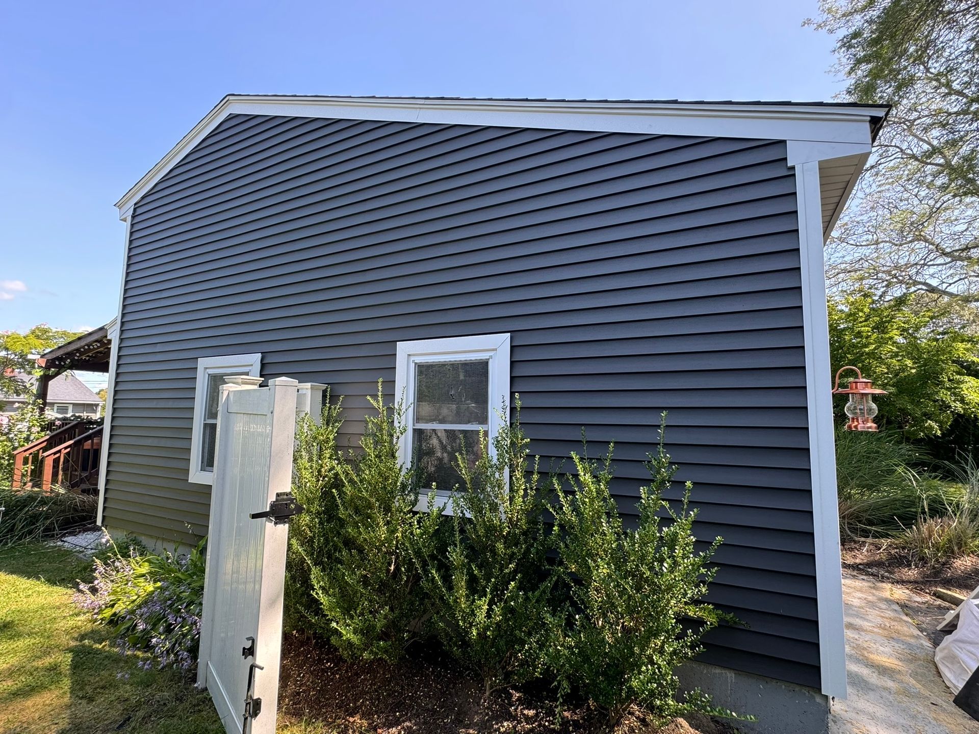 Gray-sided house exterior with white trim and bushes in front under a blue sky