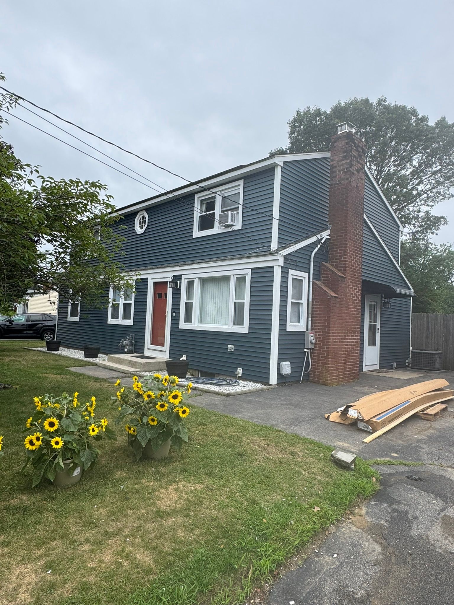 Blue-gray two-story house with red door, white trim, and a brick chimney beside a grassy yard and sunflowers